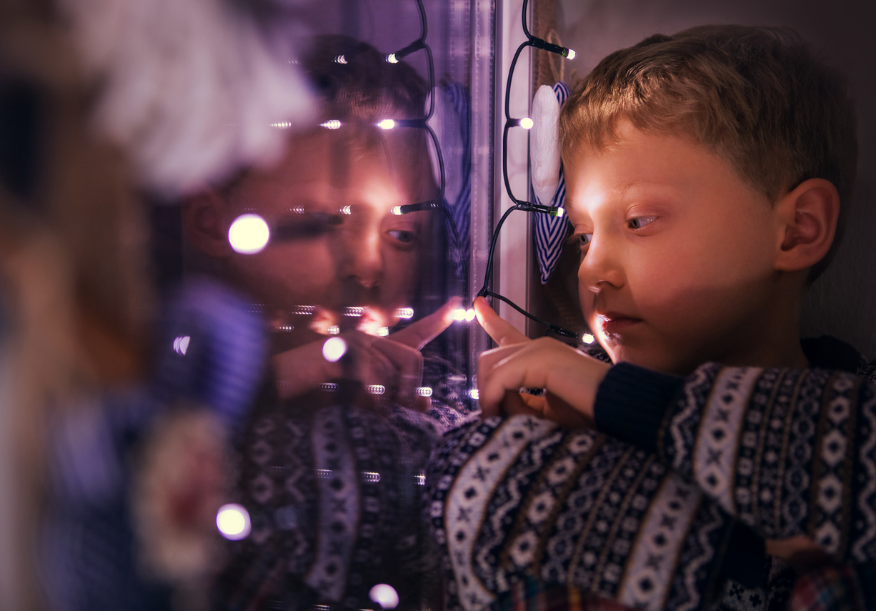 Szomorú kisfiú néz ki egy ünnepi fényekkel díszített ablakon – az ünnepi magány szimbóluma